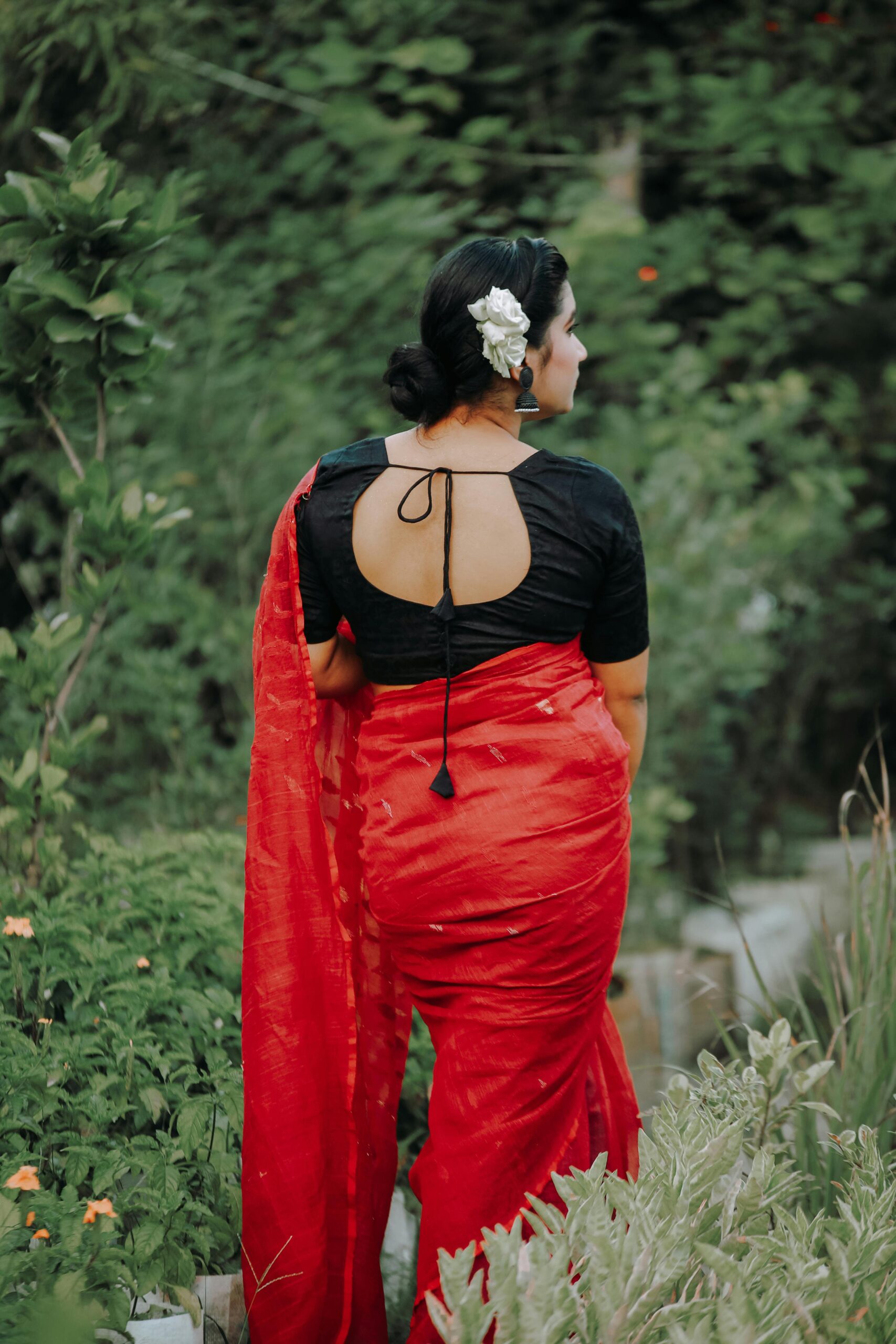 Back view of a woman in a red sari with floral hair accessory in a lush garden.